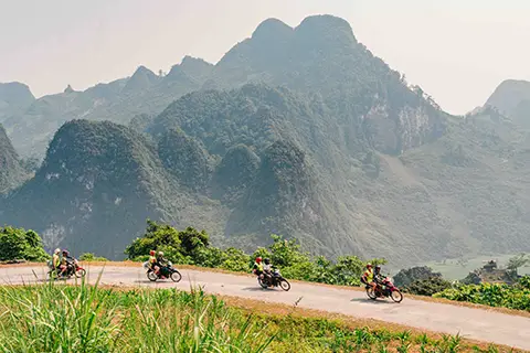 Motards sur une route de montagne avec pics karstiques et vallée brumeuse.