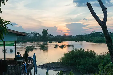 Coucher de soleil sur un large fleuve avec barques et silhouettes d’arbres.