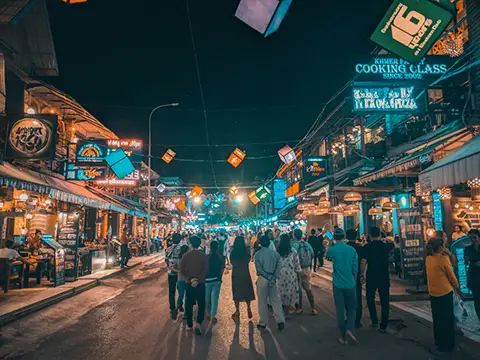 Rue de marché nocturne avec enseignes lumineuses et foule de passants.